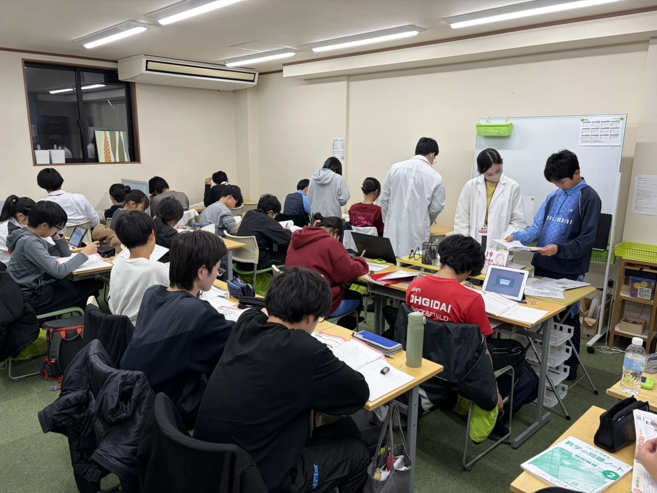 個個塾の教室で先生と生徒が教材を確認し、周囲の生徒が学習する様子